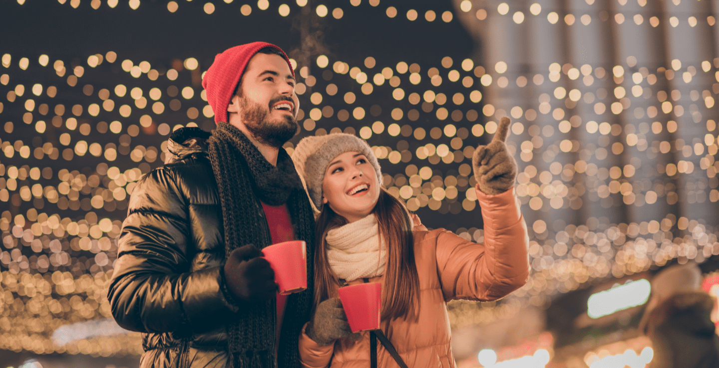 Young couple admires lights while holding hot drinks - Christmas holiday ideas